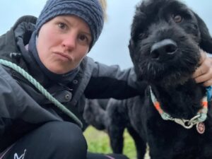 Linda kniet sich auf den Boden und umarmt einen schwarzen Hund, beide schauen in die Kamera