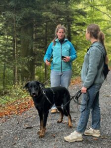 Einzeltraining im Wald: Linda erklärt einer Frau etwas, die Frau schaut Linda an. Ihr Hund steht neben ihr an der Leine und schaut in die Kamera