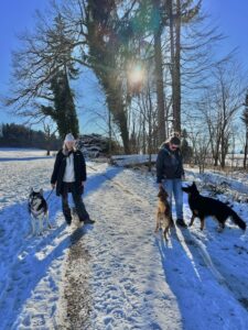3 Hunde, 2 Menschen stehen im Schnee auf einem Weg und schauen in die Kamera