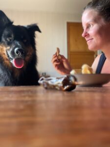 Hundeernährung: Linda sitzt am Tisch und hat etwas in der Hand, ein Kauknochen liegt auf dem Tisch. Akay schaut auf den Kauknochen
